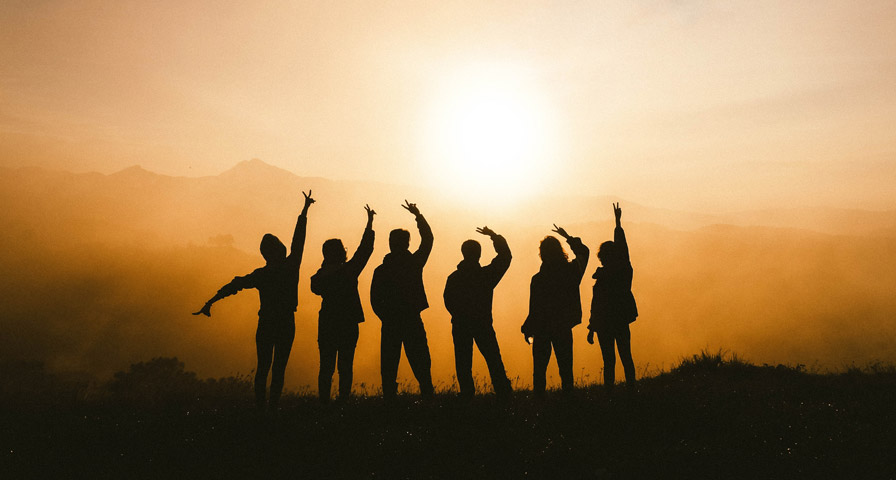 Sillouette of six people raising arms in celebration in front of a sunrise.